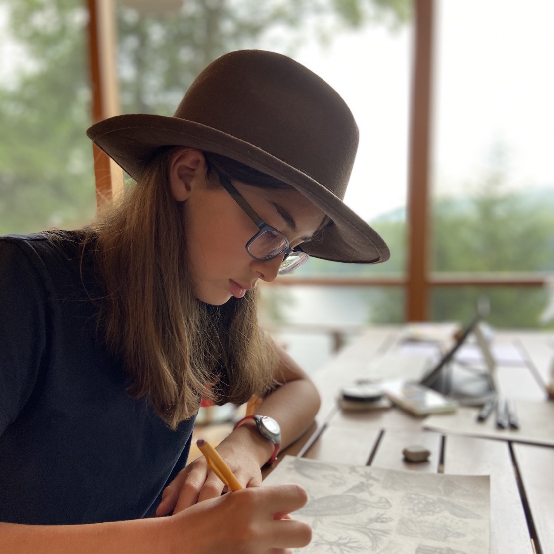 A picture of Frances. Frances is wearing a fedora and a dark blue shirt and she is drawing a picture.